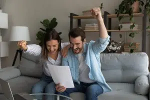 excited married couple hug couch yell yes read paper letter about getting tax refund money grant family business amazed young spouses overjoyed by perfect conditions loan mortgage agreement Digital Ware